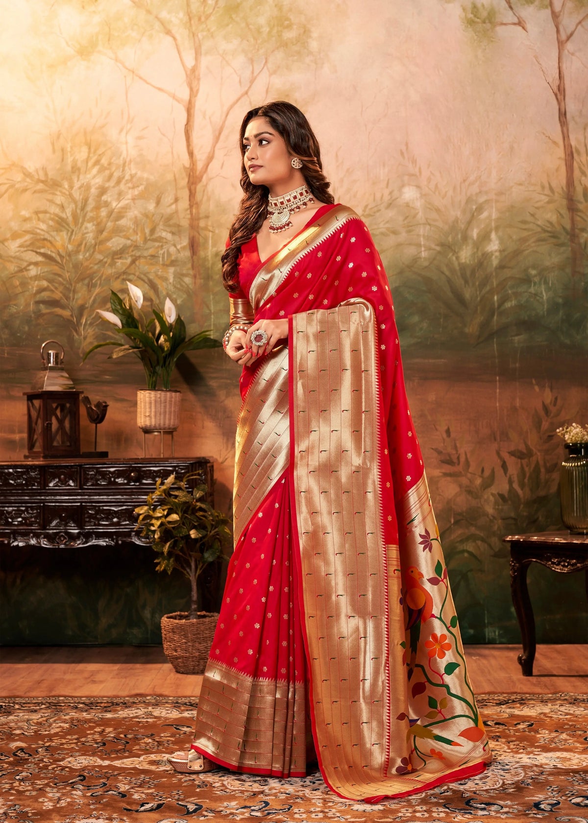 Woman in a red and gold saree standing in a decorated room with plants and furniture.