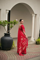 Woman in a red saree standing in an outdoor setting with plants and columns.