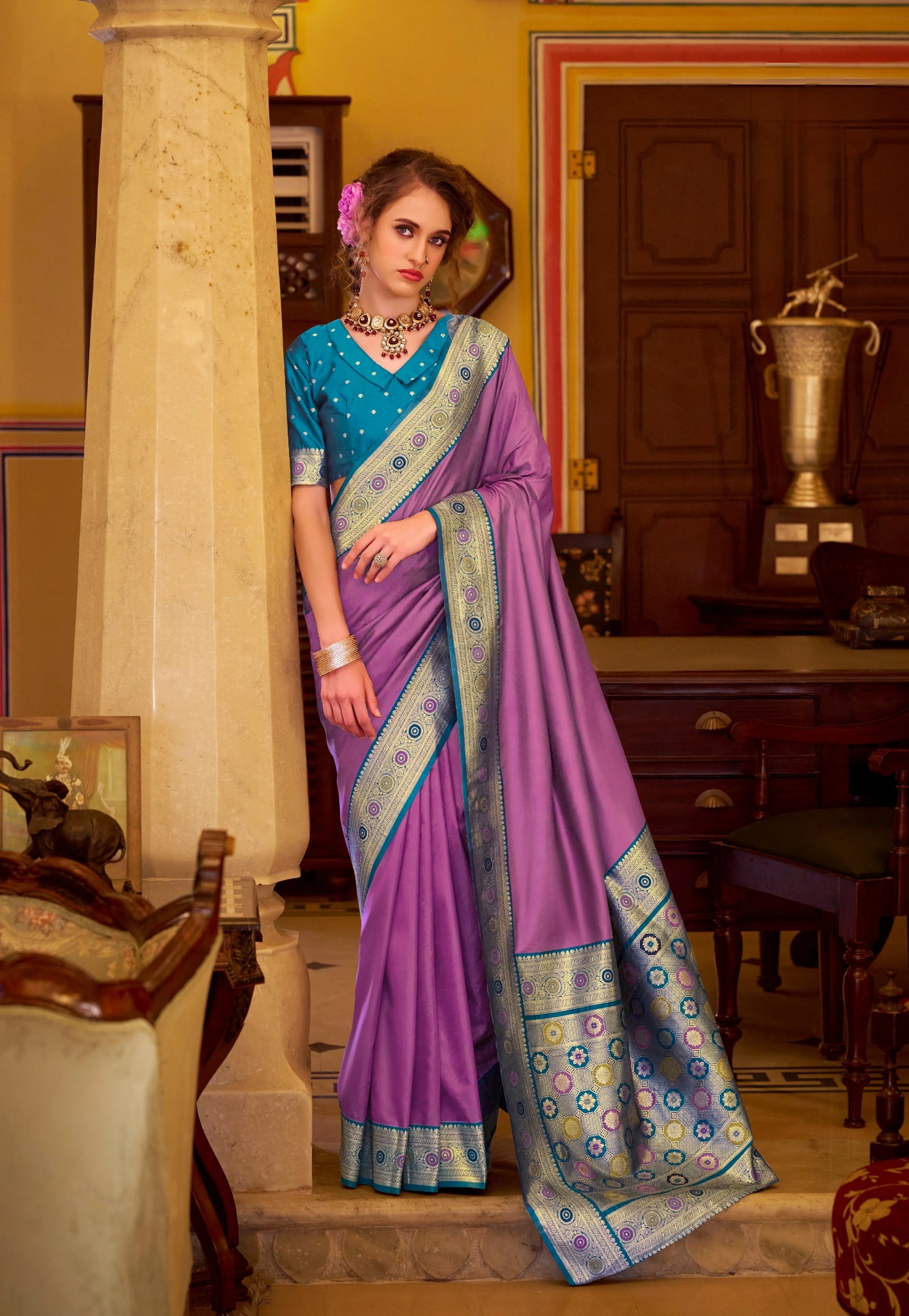 Woman in a blue and purple saree standing in an indoor setting with trophies and furniture.