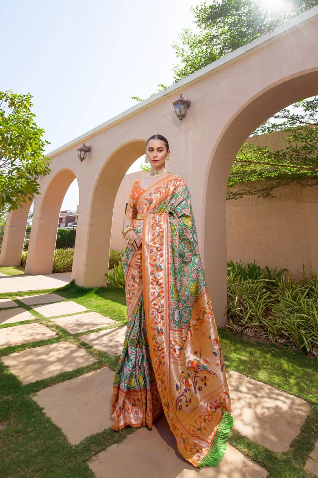 Woman in a colorful saree standing in a garden with arches and greenery.