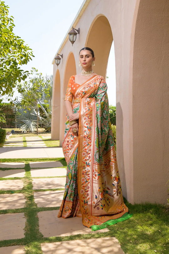 Woman in a colorful saree standing outdoors near a building with greenery.