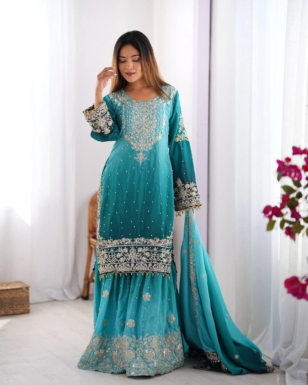 Woman wearing a teal embroidered traditional outfit with a white curtain and flowers in the background