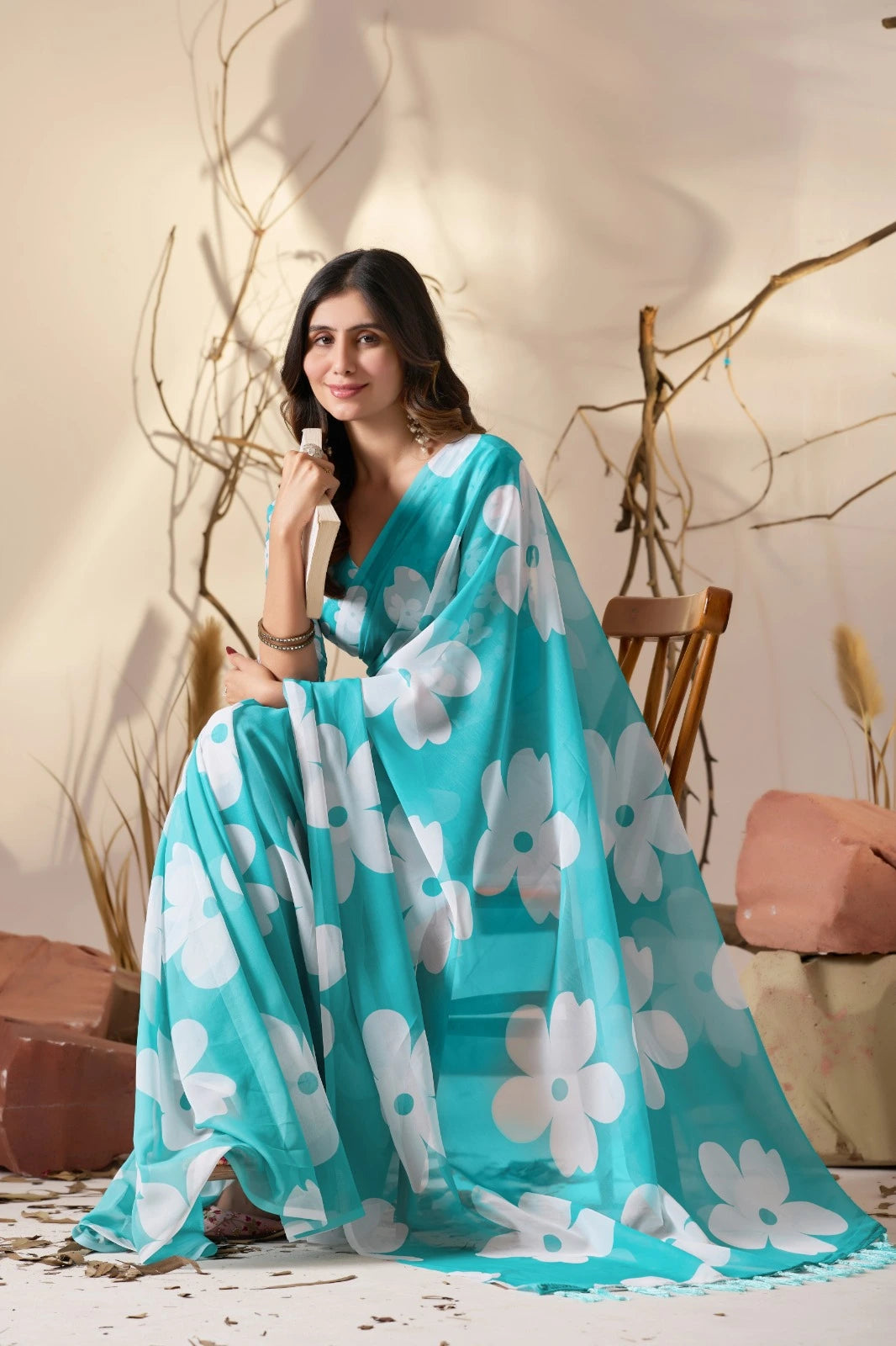 Woman wearing a turquoise saree with white floral patterns, sitting indoors.