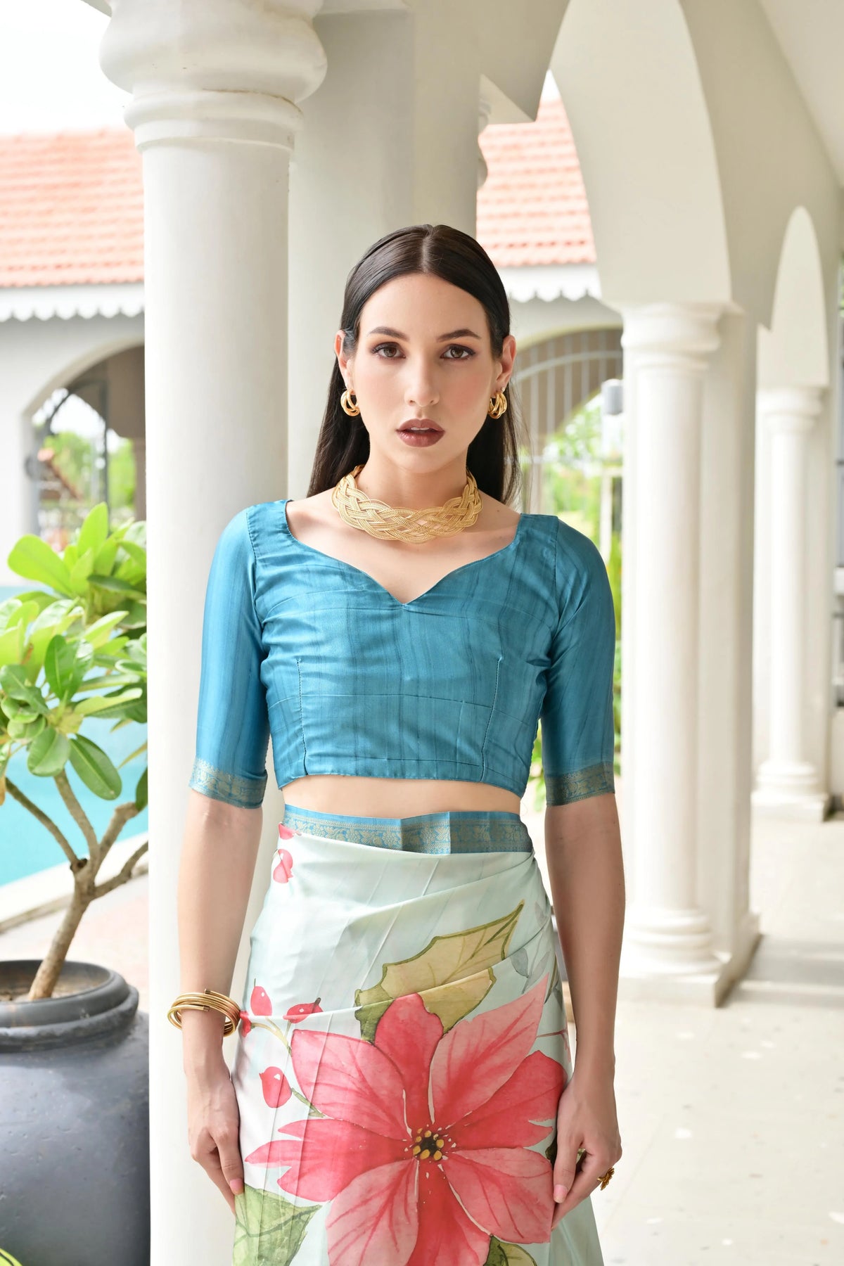 Woman wearing a blue blouse and floral skirt in an outdoor setting with columns and plants.