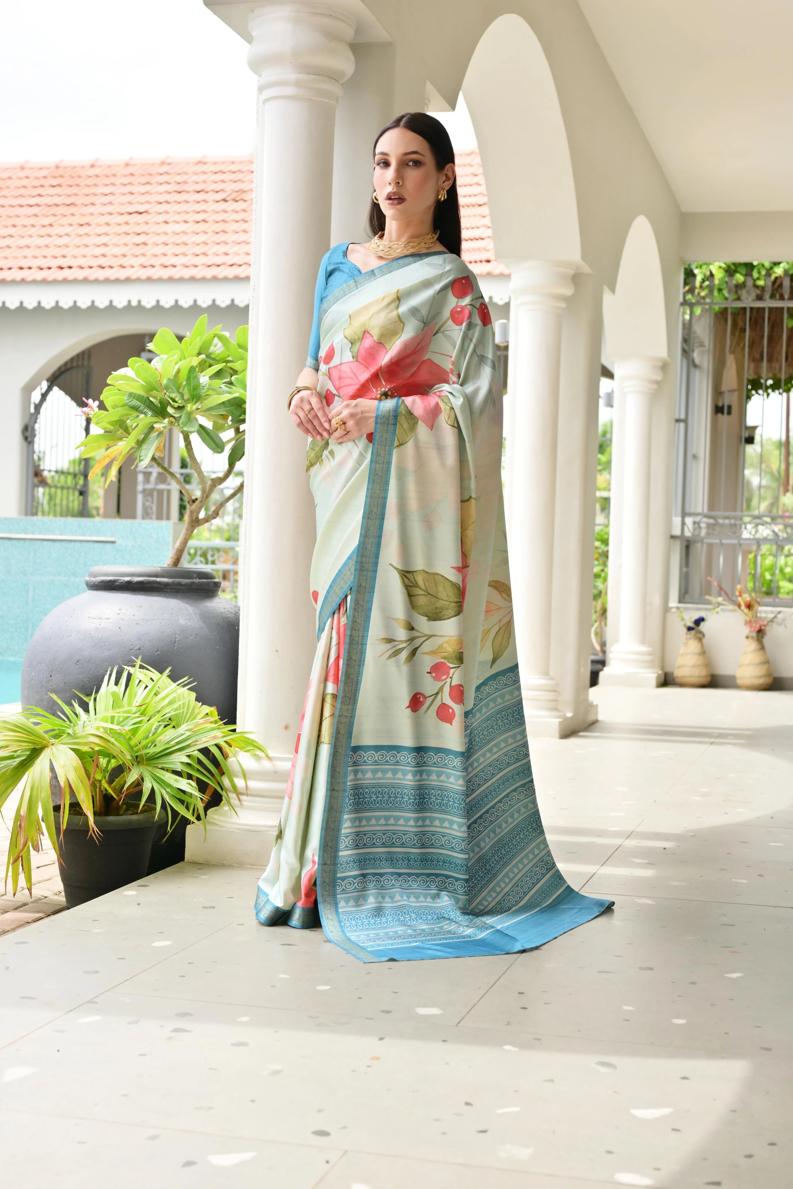Woman wearing a floral saree standing on a porch with plants and architectural elements in the background.