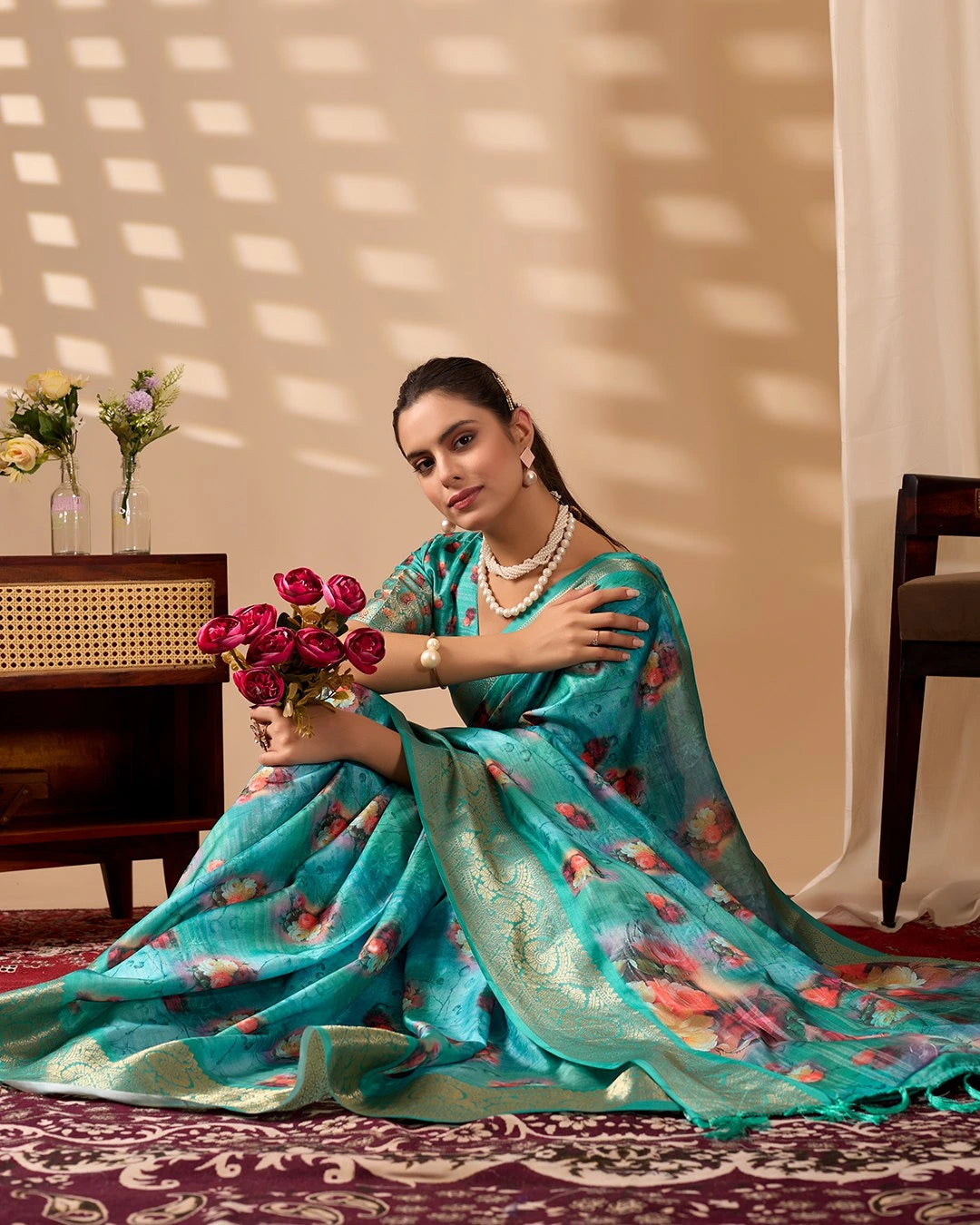 Woman in a turquoise saree with floral patterns sitting indoors, holding flowers.