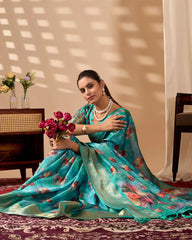 Woman in a turquoise saree with floral patterns sitting indoors, holding flowers.