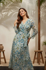 Woman wearing a floral dress standing in a room with wooden stools and plants.