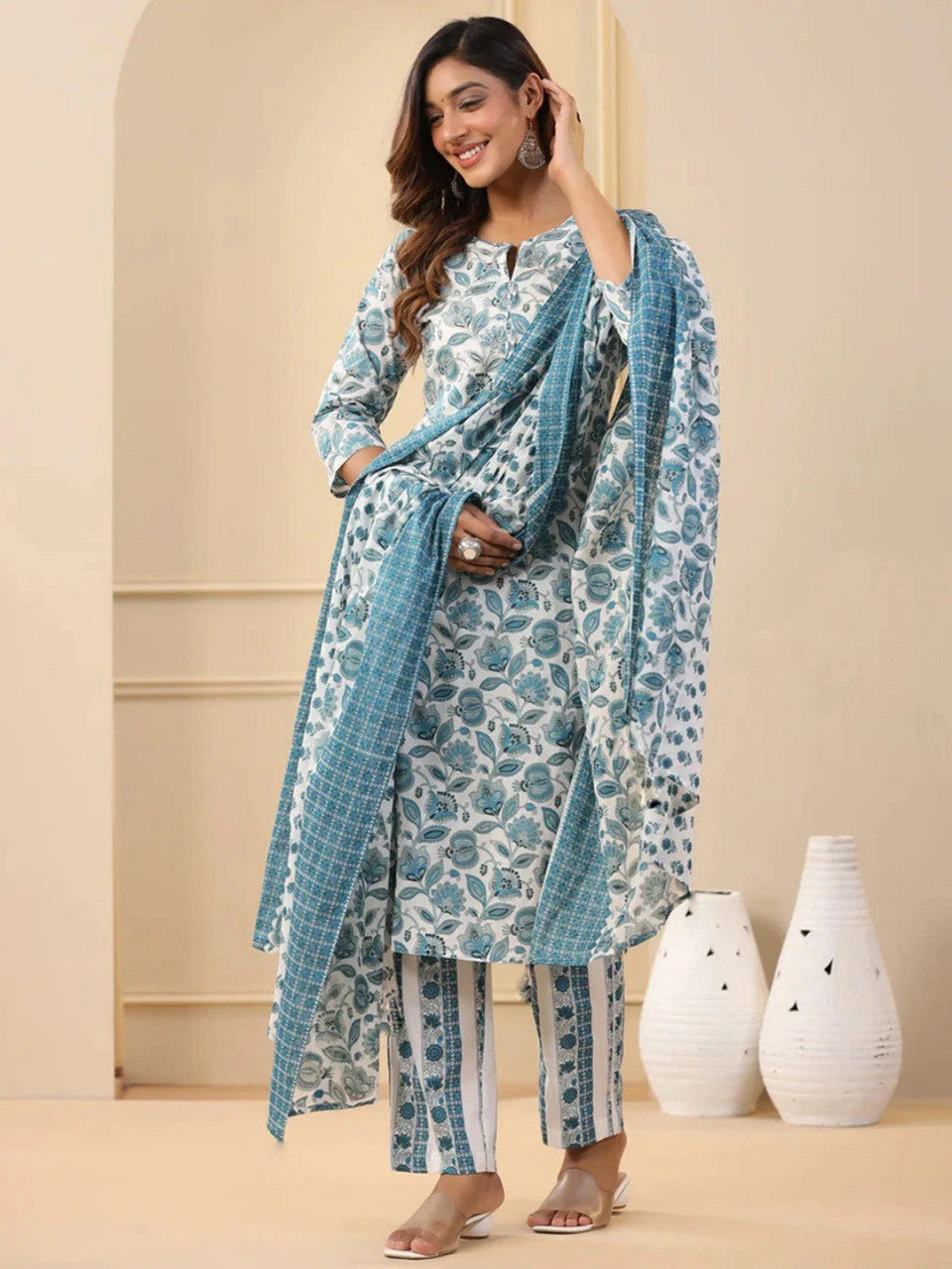 Woman wearing a blue and white patterned outfit with a scarf, standing against a beige wall with decorative vases.