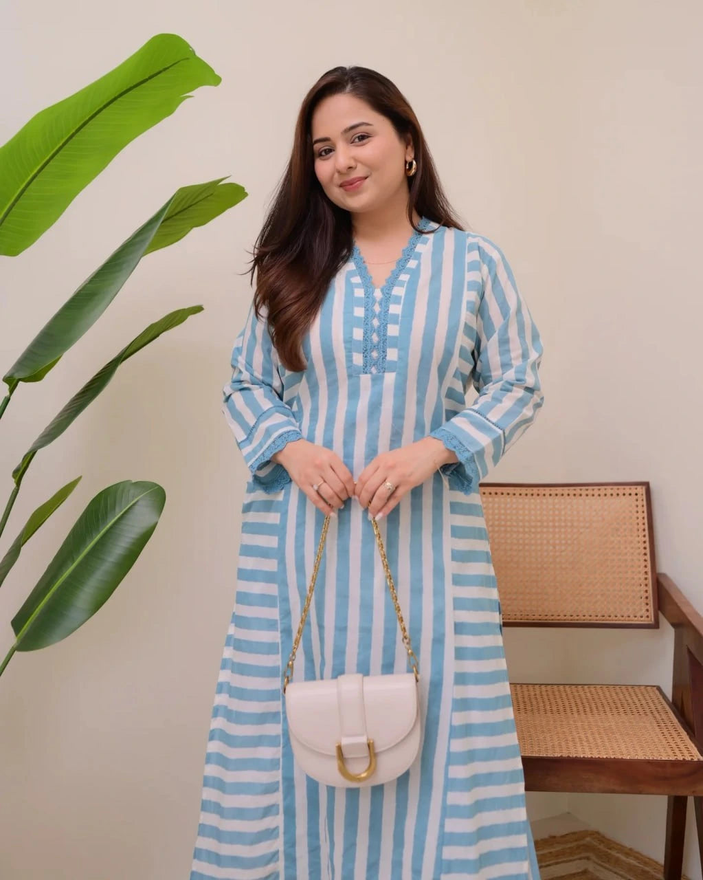Woman in a blue and white striped dress holding a white handbag indoors with a plant and chair in the background.