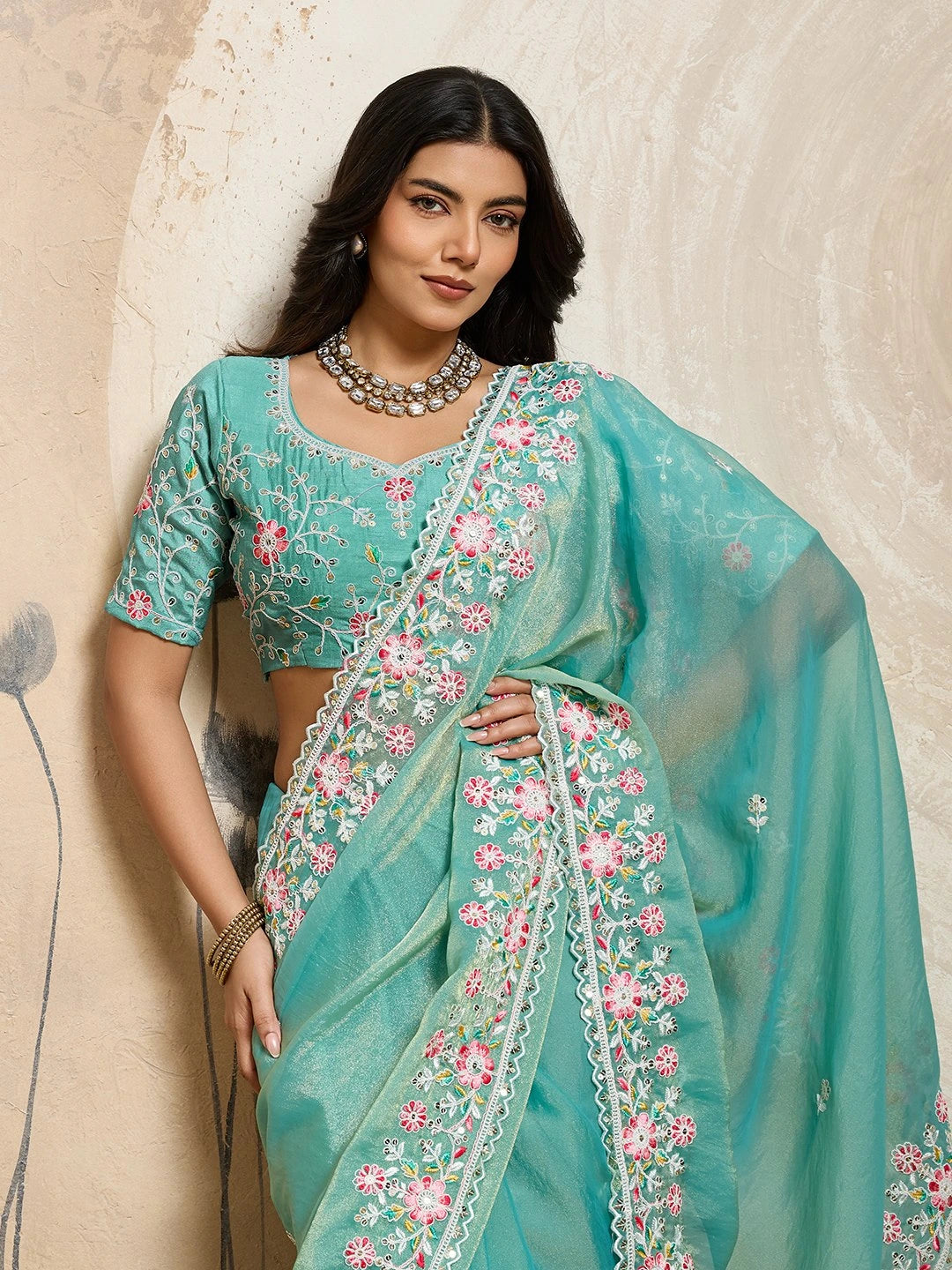 Woman wearing a teal saree with floral patterns against a beige wall.