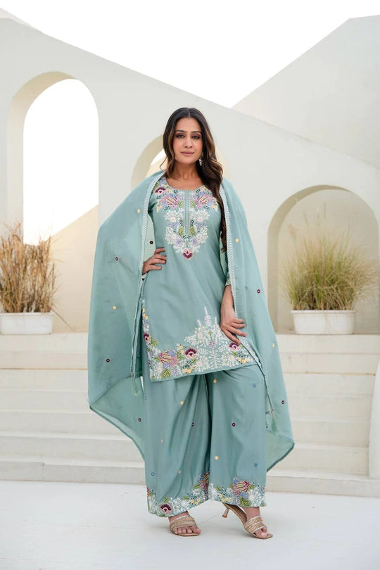 Woman in a light blue embroidered traditional outfit standing in a decorative setting.