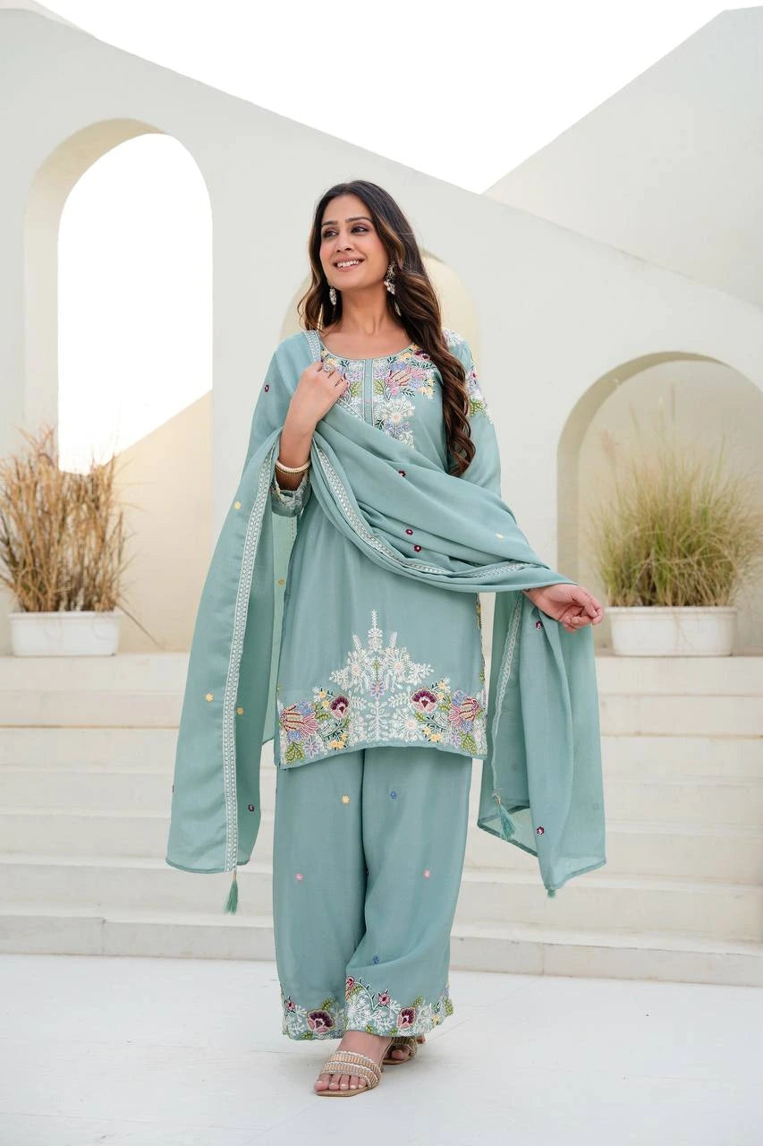 Woman in a light blue traditional outfit with floral embroidery standing in a decorative outdoor setting.