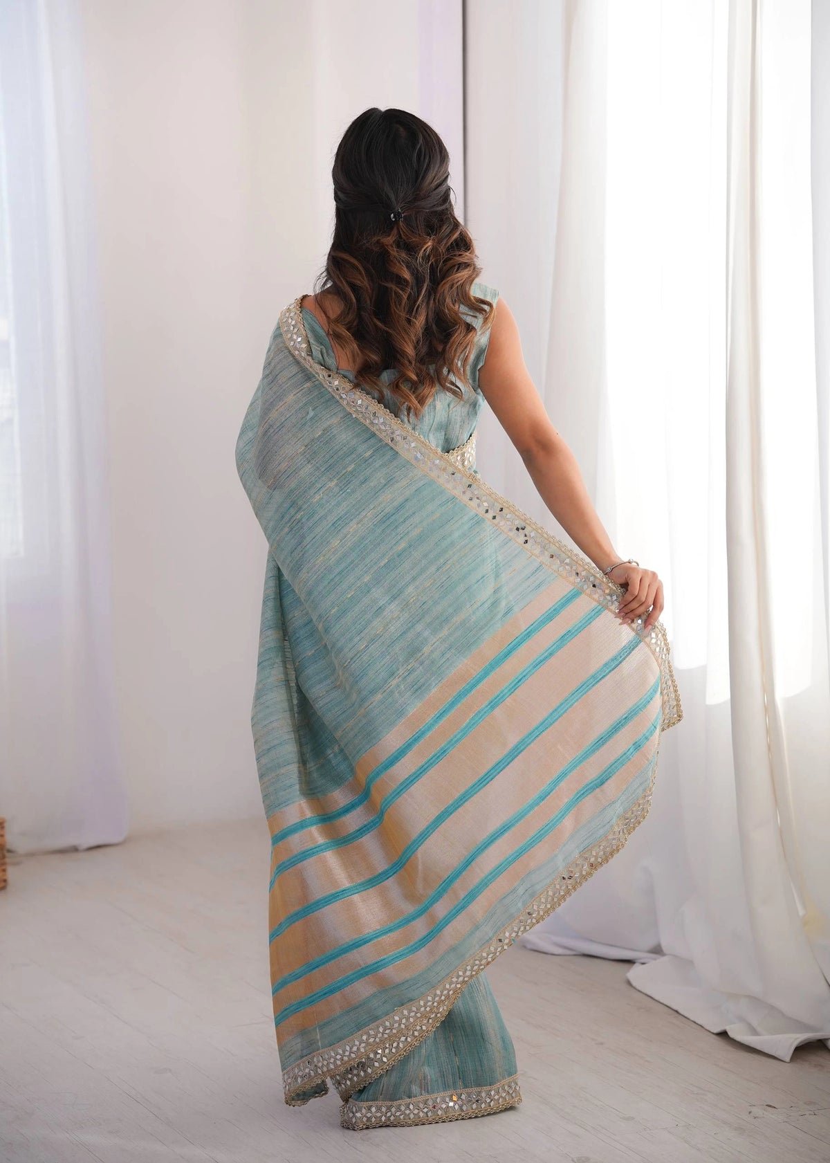 Woman wearing a light blue and beige striped saree in a room with white curtains.