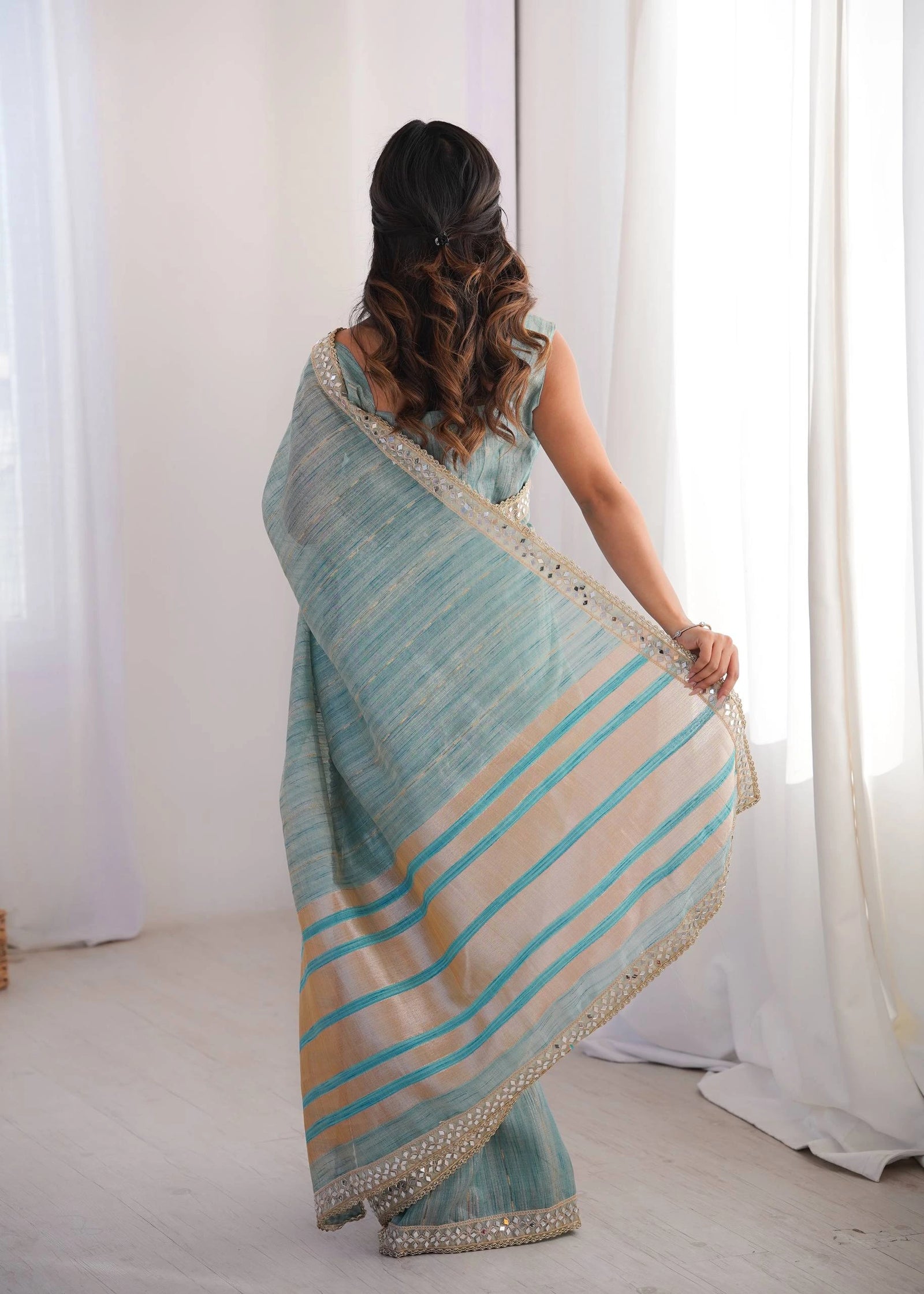 Woman wearing a light blue and beige striped saree in a room with white curtains.