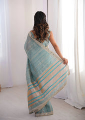 Woman wearing a light blue and beige striped saree in a room with white curtains.