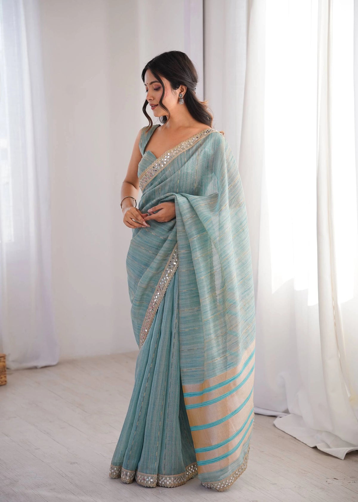 Woman wearing a light blue saree with white and gold border against a white curtain background
