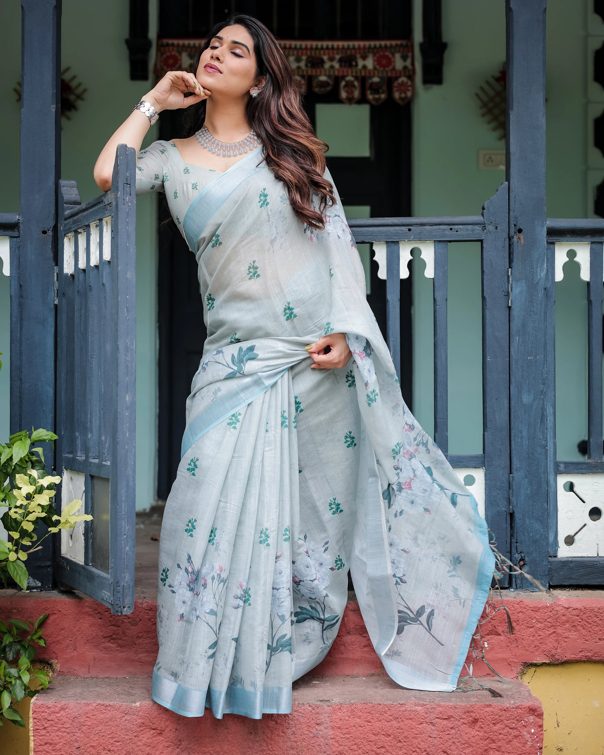 Woman in a light blue saree with floral patterns standing on a porch.