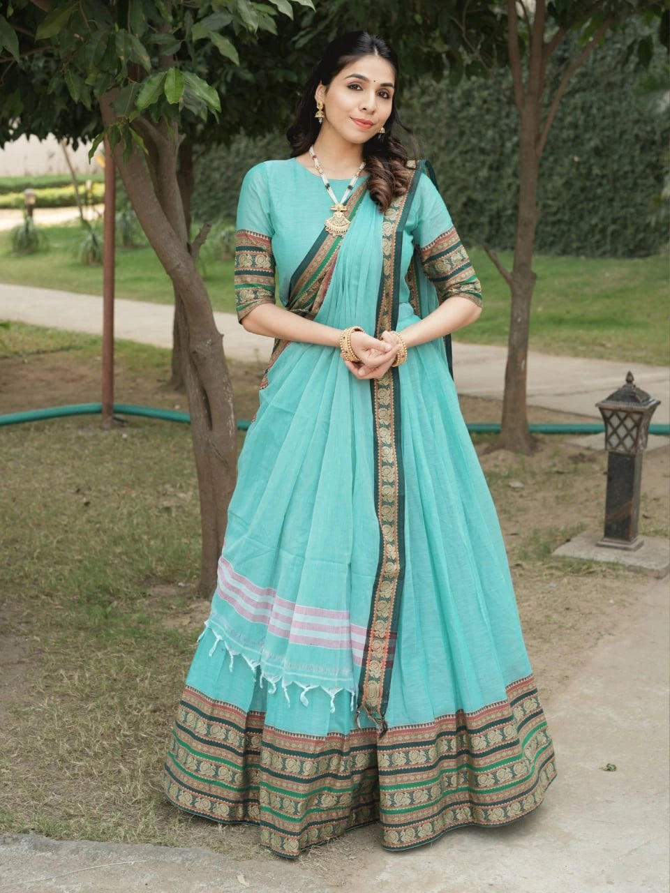 Woman wearing a light blue saree with green and brown patterns in an outdoor setting.