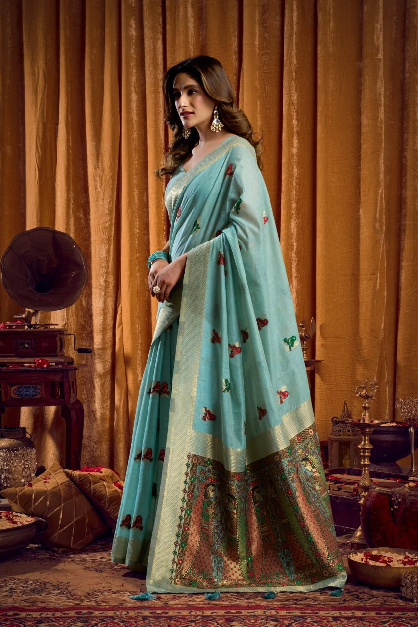 Woman in a light blue saree with floral patterns standing in front of a decorative background.