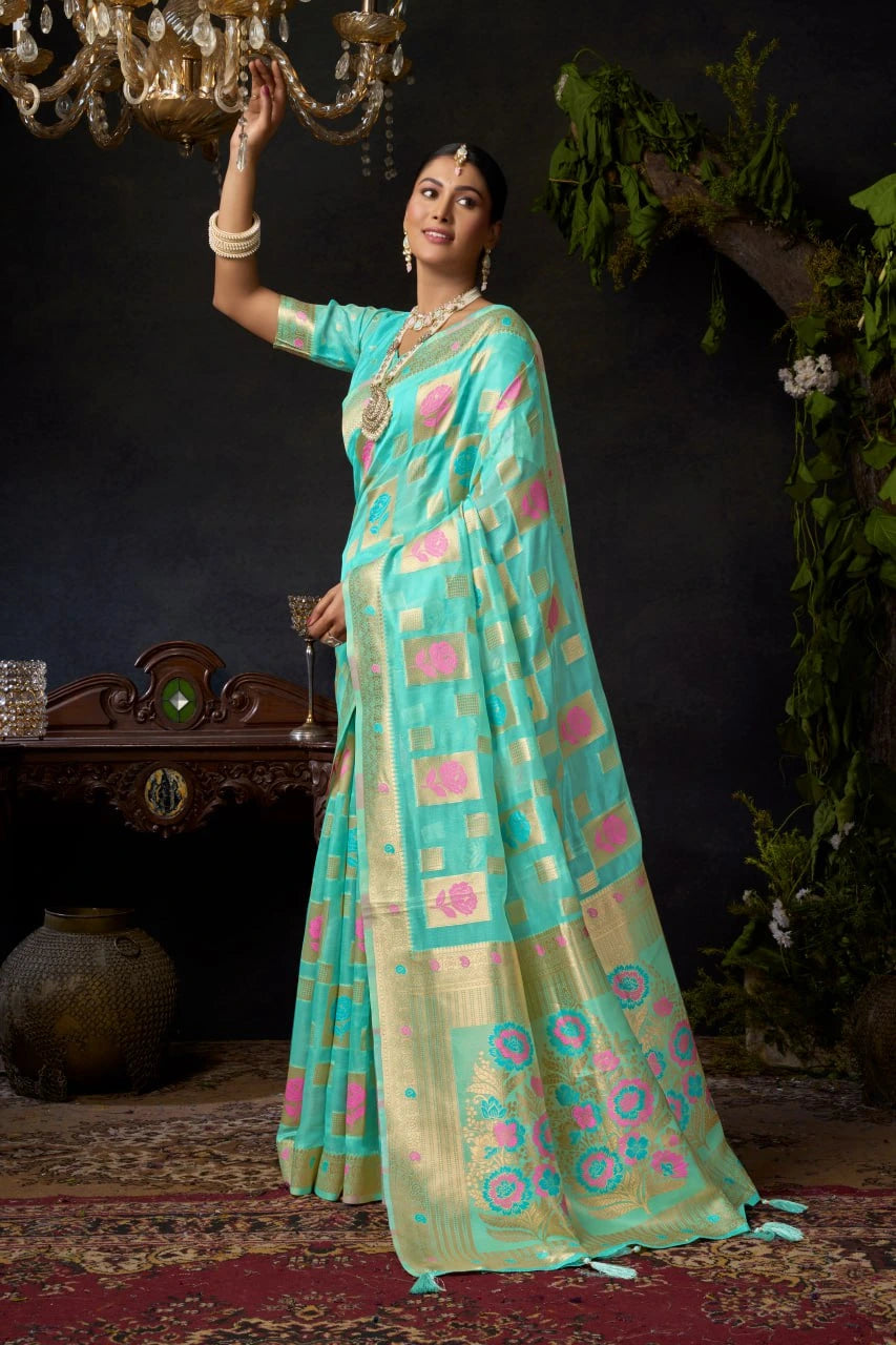 Woman in a traditional turquoise saree with pink patterns, standing indoors with decorative background.