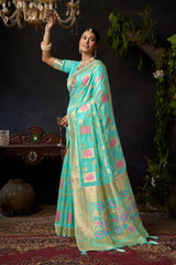 Woman in a traditional turquoise saree with pink patterns, standing indoors with decorative background.