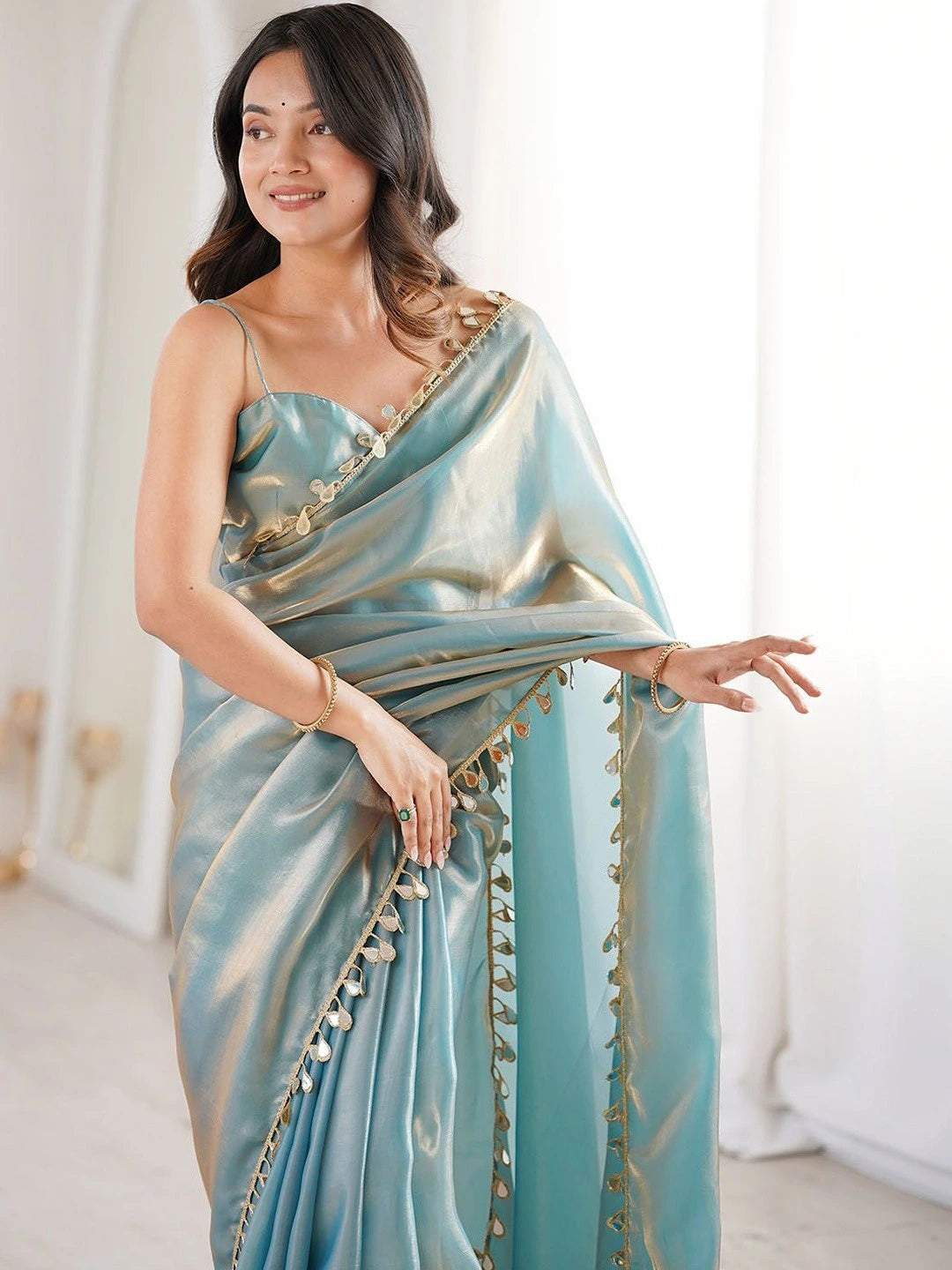 Woman wearing a light blue saree with gold accents in a bright room.