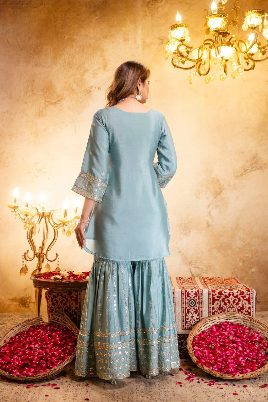 Woman in a light blue traditional outfit standing in a decorated room with chandeliers and red flowers.
