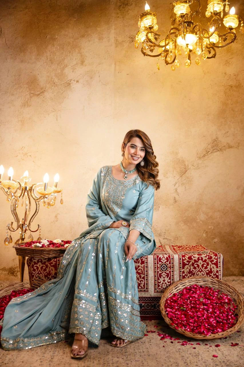 Woman in a light blue traditional outfit sitting on a decorative chair with a chandelier and rose petals in the background.