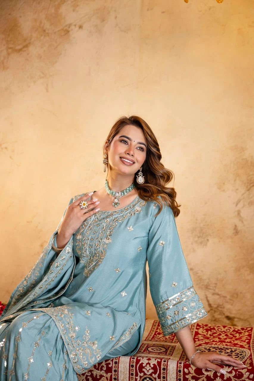 Woman in a light blue embroidered traditional outfit sitting on a patterned rug against a beige wall.