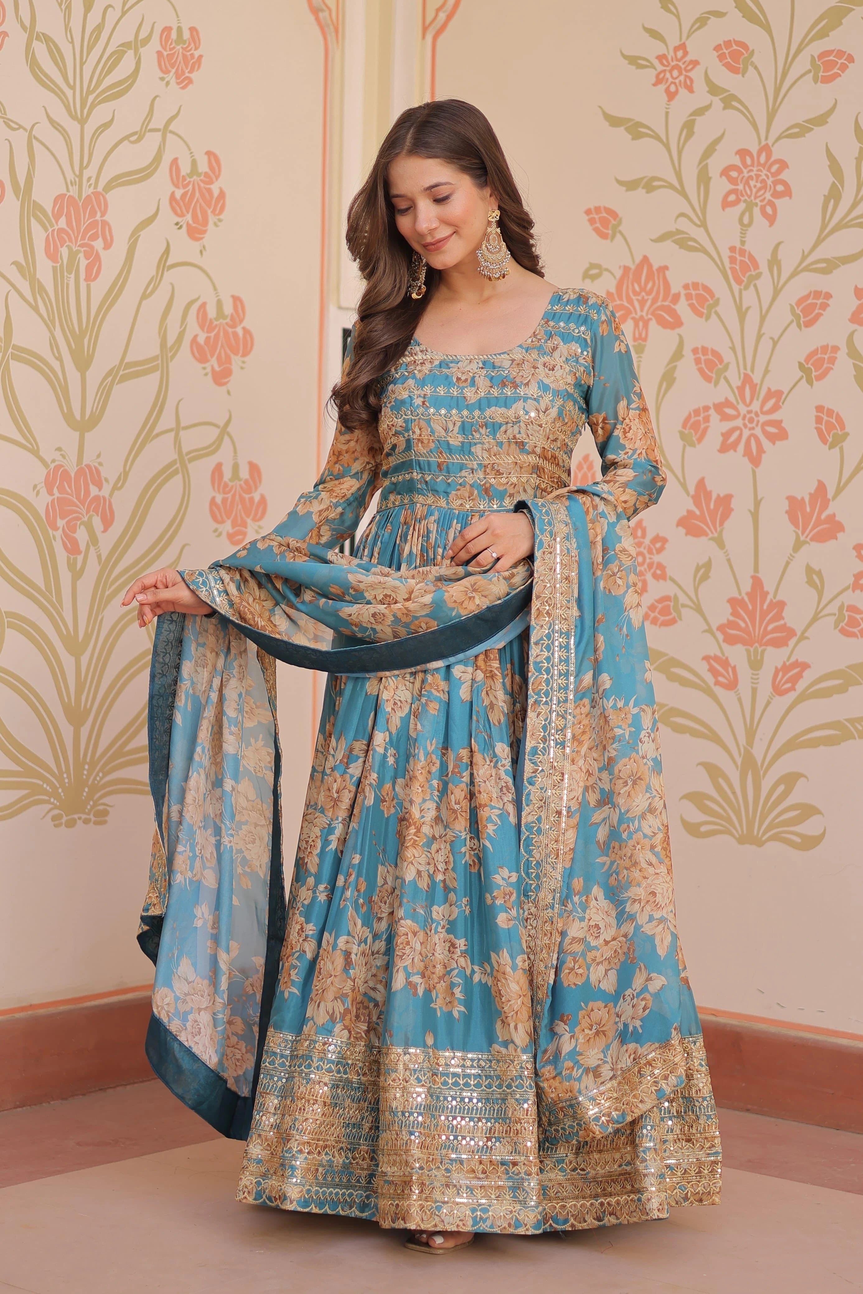 Woman wearing a blue and gold floral dress with a matching dupatta against a decorative wall.