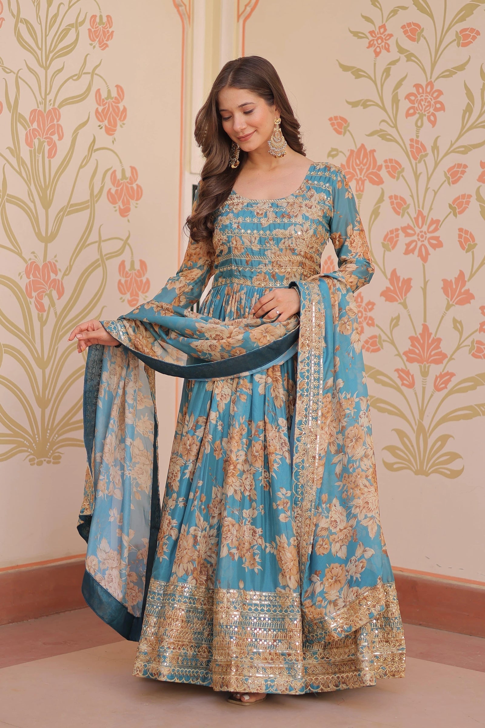 Woman wearing a blue and gold floral dress with a matching dupatta against a decorative wall.
