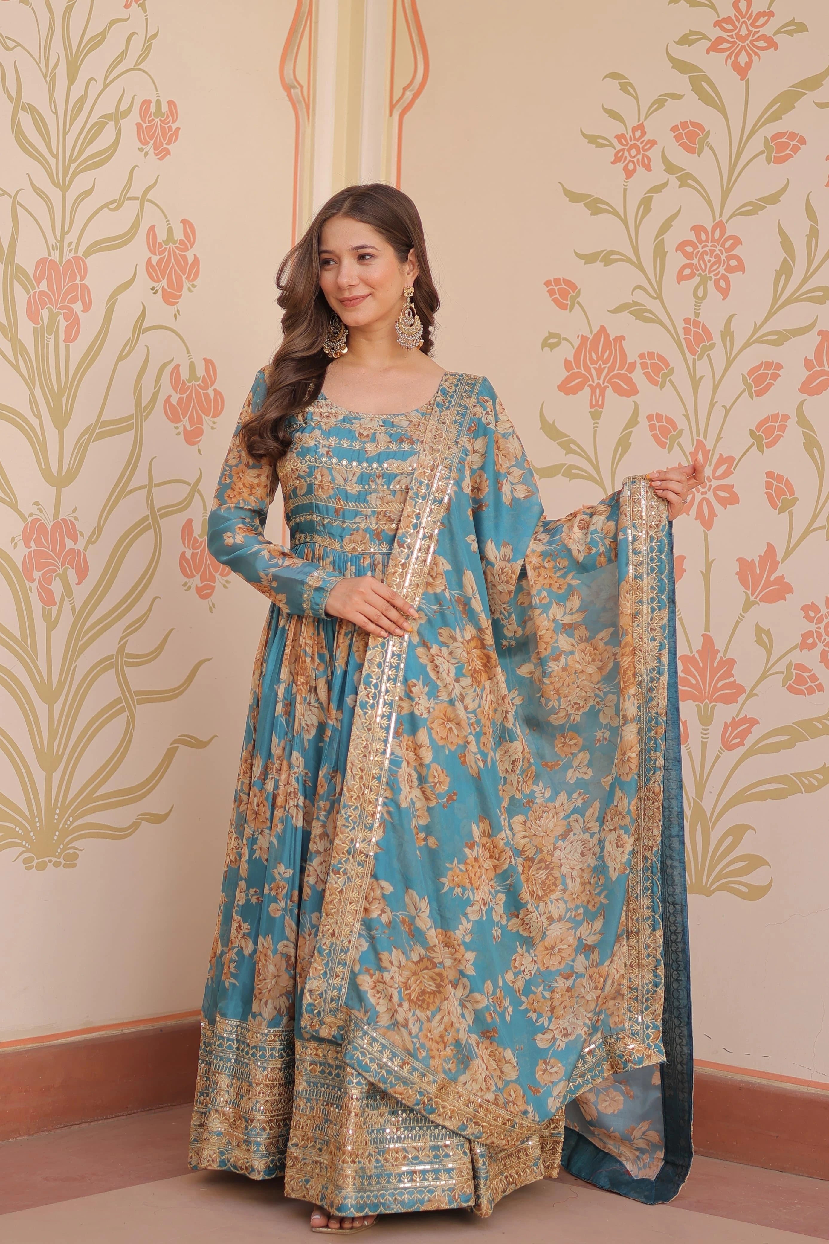 Woman in a blue and gold traditional outfit with a floral pattern against a decorative wall.