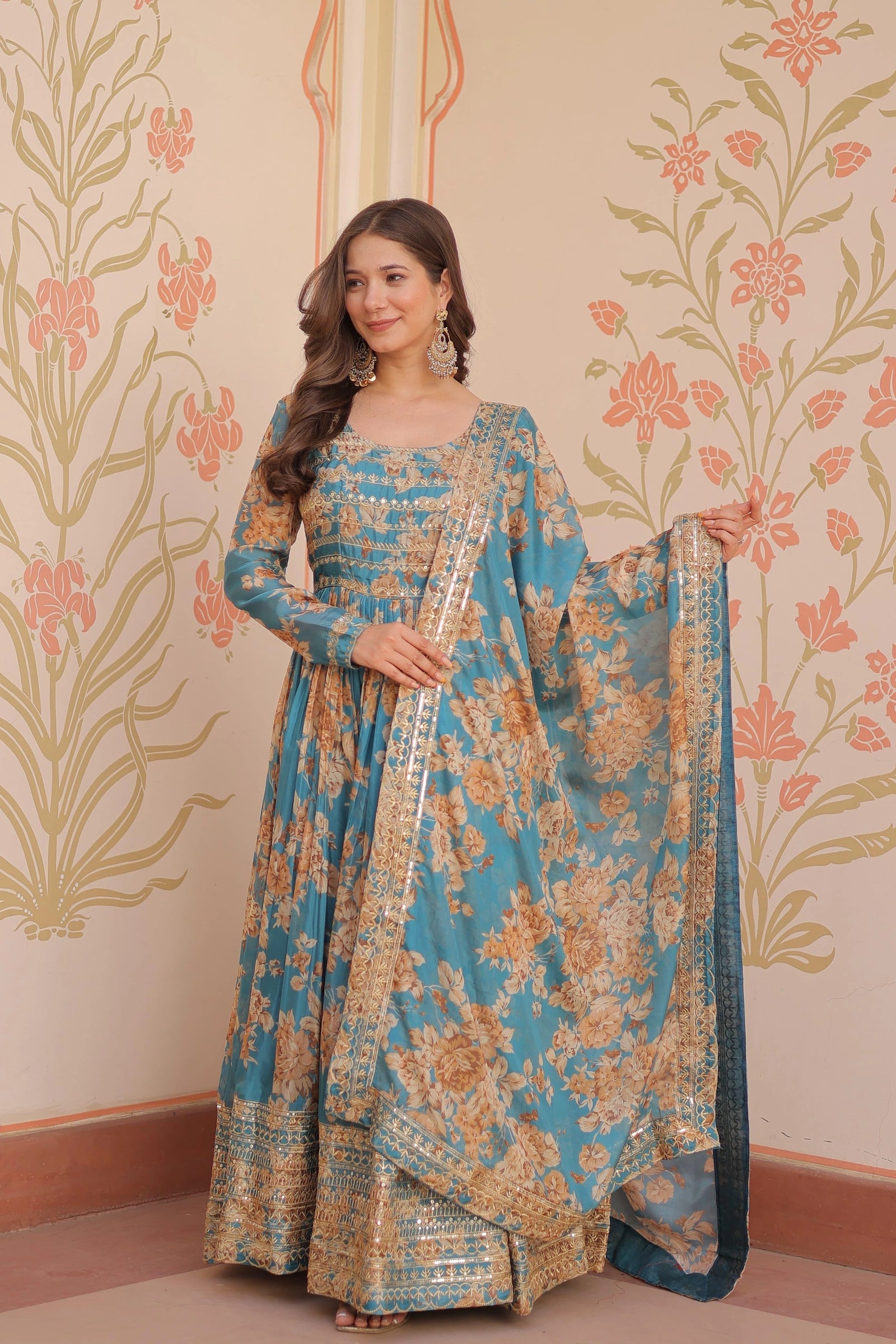 Woman in a blue and gold traditional outfit with a floral pattern against a decorative wall.