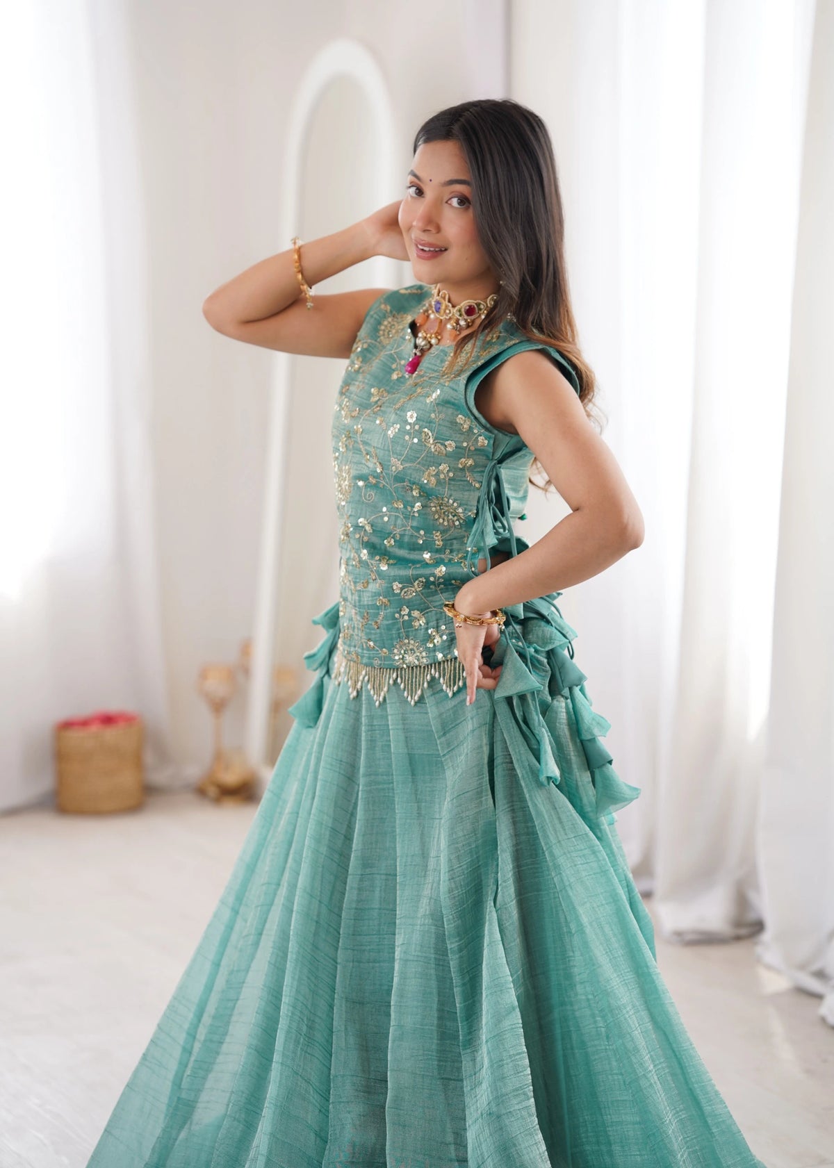 Woman in a teal dress with gold embellishments posing in a bright room.