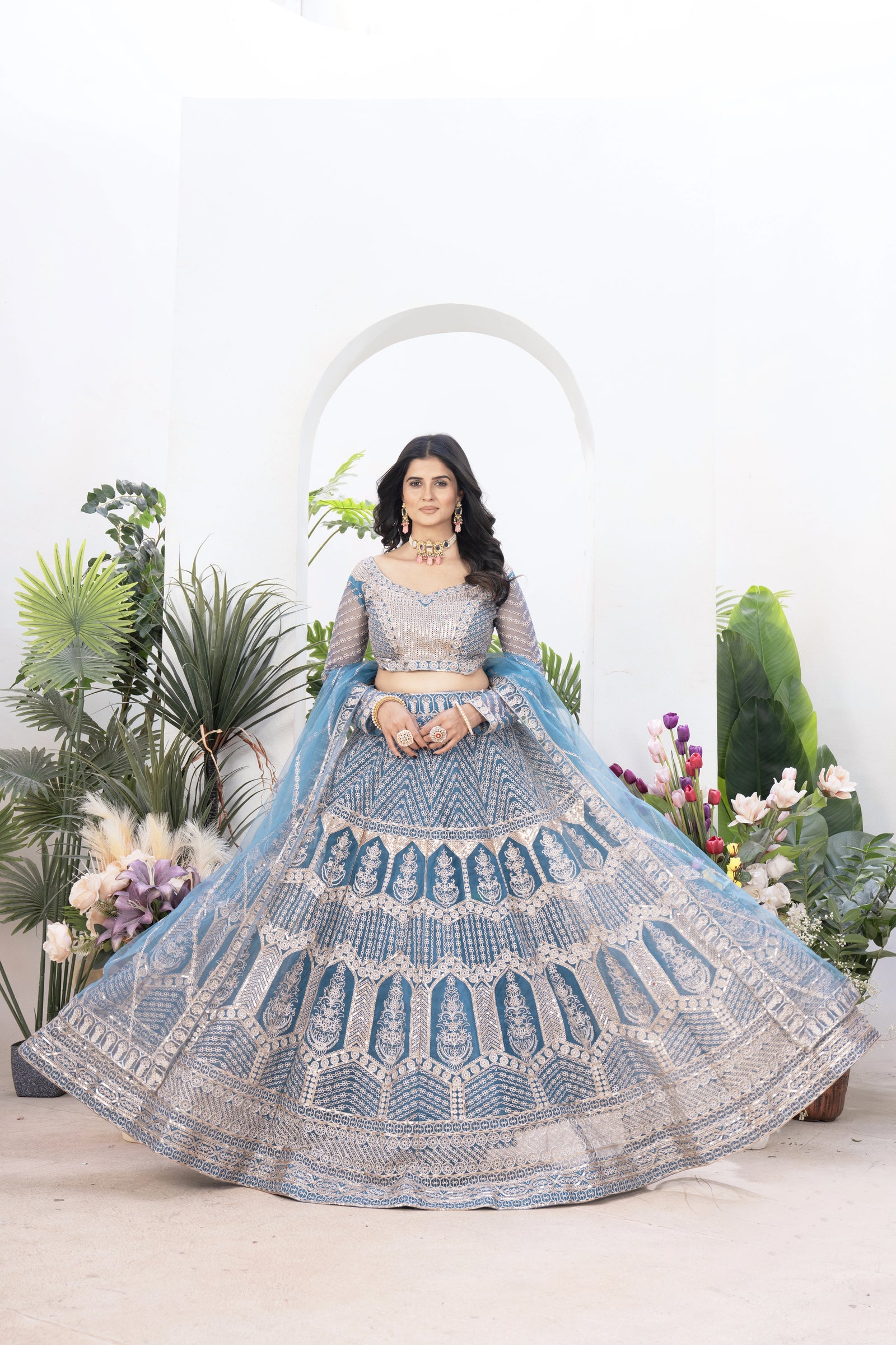 Woman in a blue embroidered traditional outfit with floral decorations and plants in the background