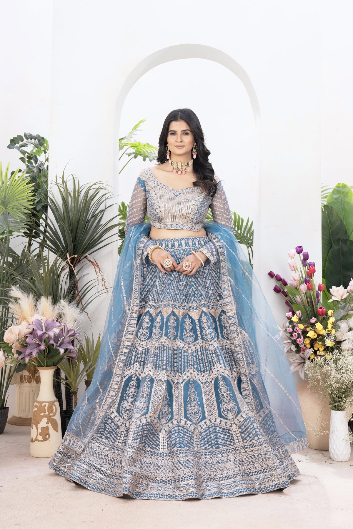 Woman in a blue and silver traditional outfit with floral decorations and plants in the background