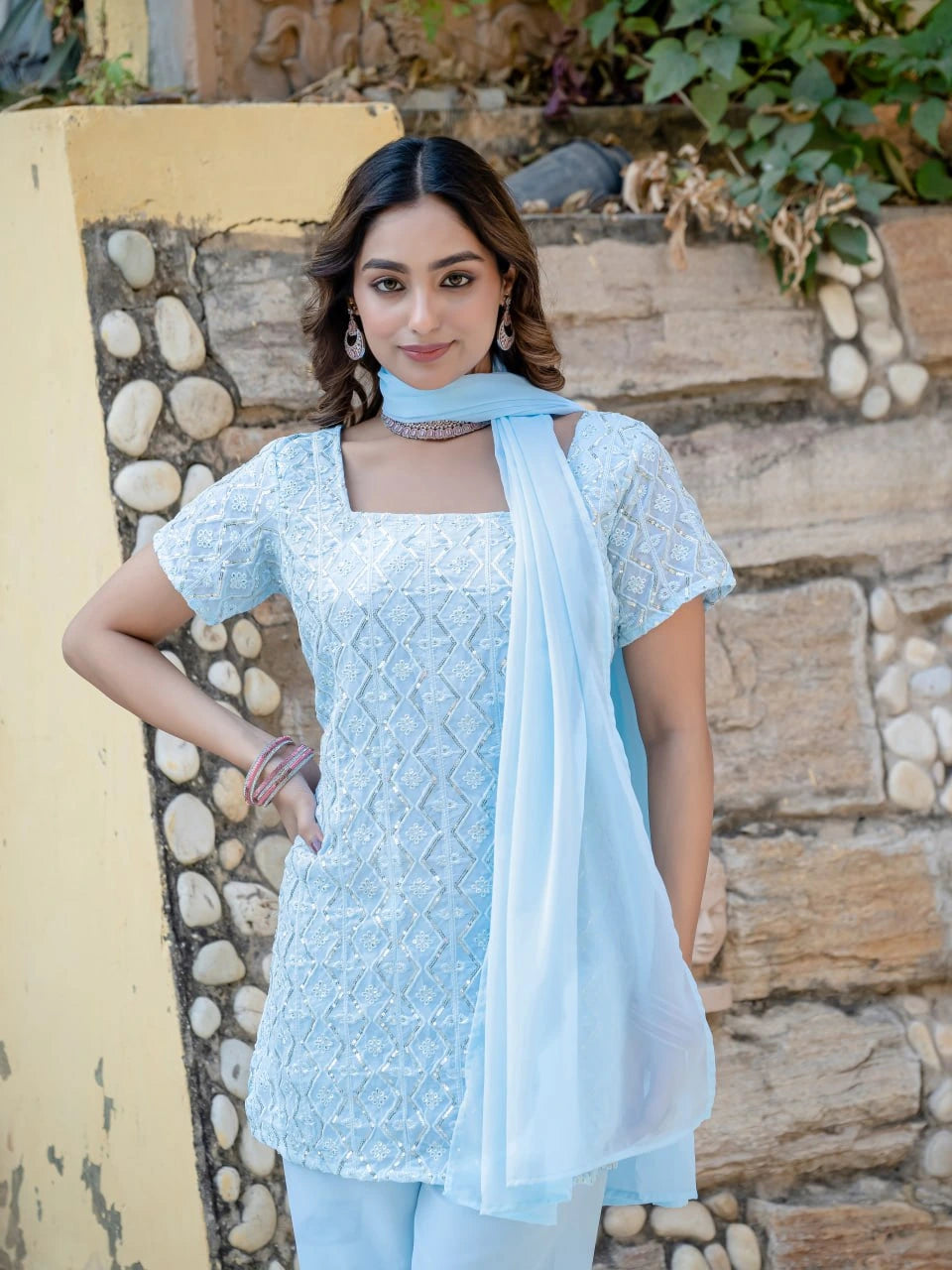 Woman wearing a light blue embroidered outfit with a scarf against a stone wall background