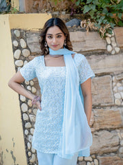 Woman wearing a light blue embroidered outfit with a scarf against a stone wall background