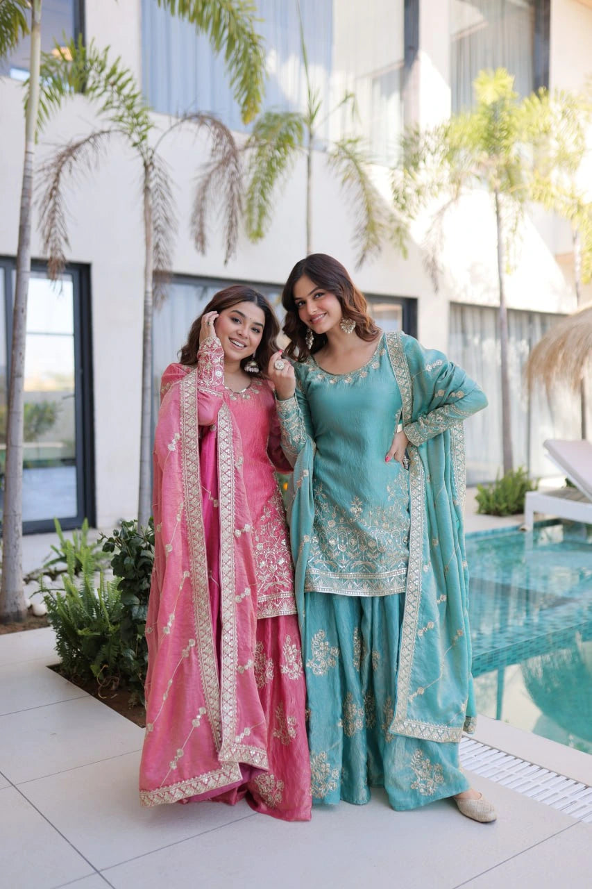 Two women in traditional outfits standing by a poolside with plants and a building in the background.