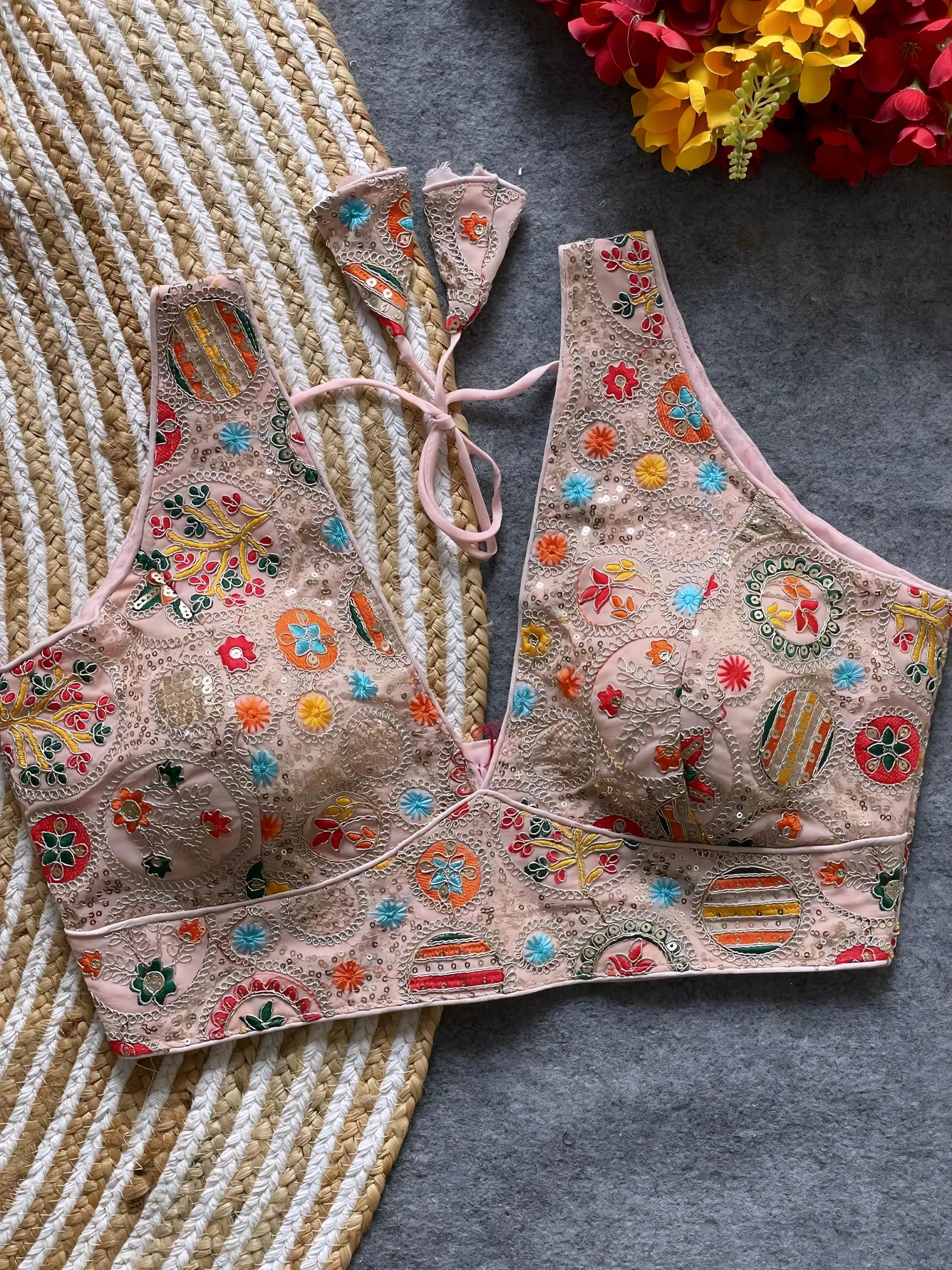 Traditional blush pink blouse featuring vibrant multicolor thread embroidery and mirror work in circular floral and geometric patterns, with sequin accents and a deep V-neck tie-back detail, displayed flat on a textured mat with artificial red and yellow flowers in the corner.