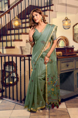 Woman in a green saree standing in an indoor setting with staircase and decor.