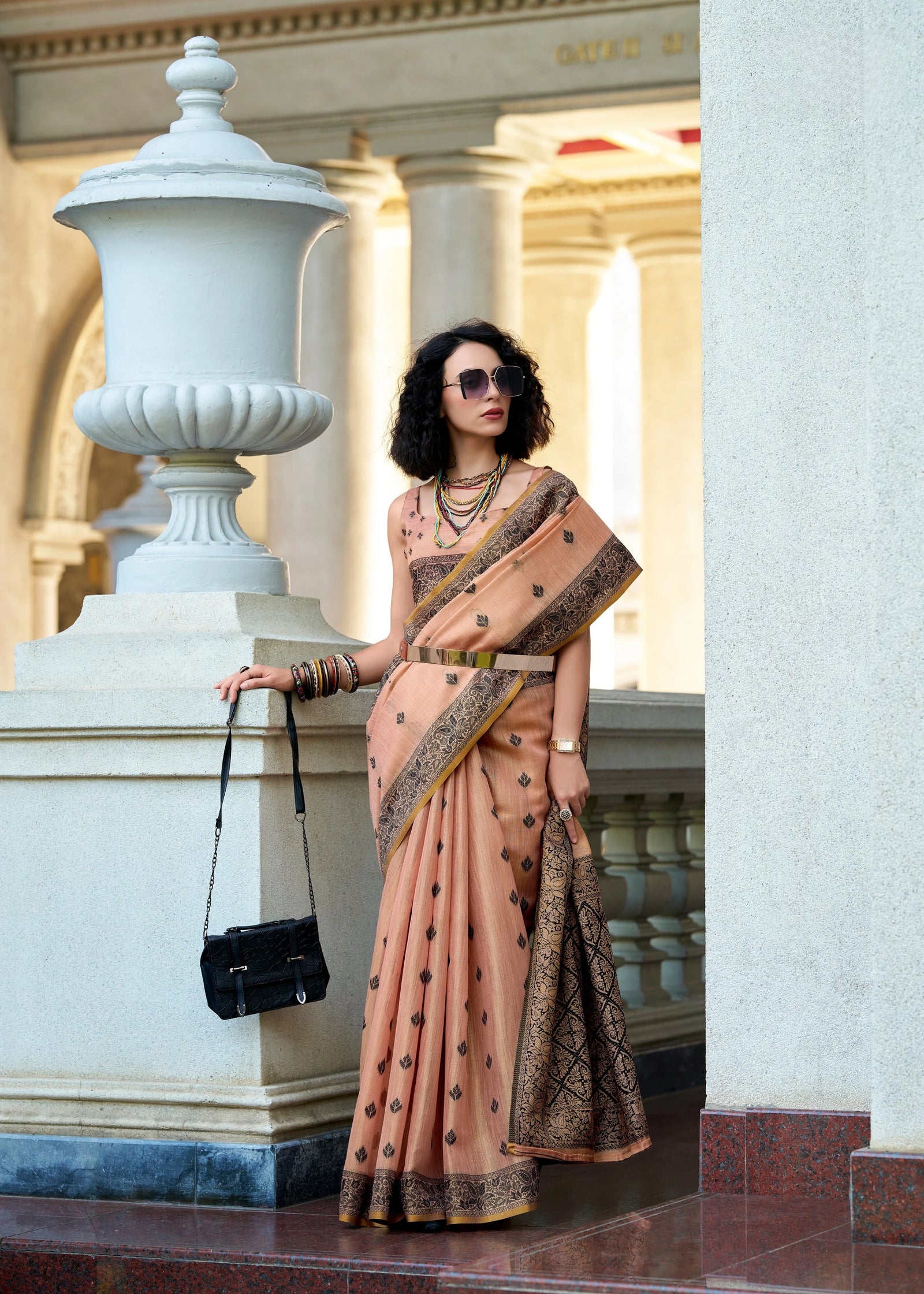 Woman in a peach saree with black patterns standing in front of classical architecture.