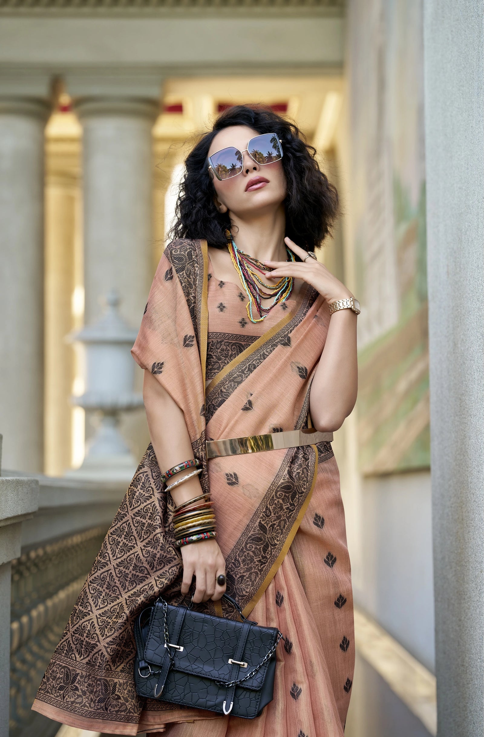 Woman in a peach saree with intricate patterns, holding a black handbag, in an elegant interior setting.