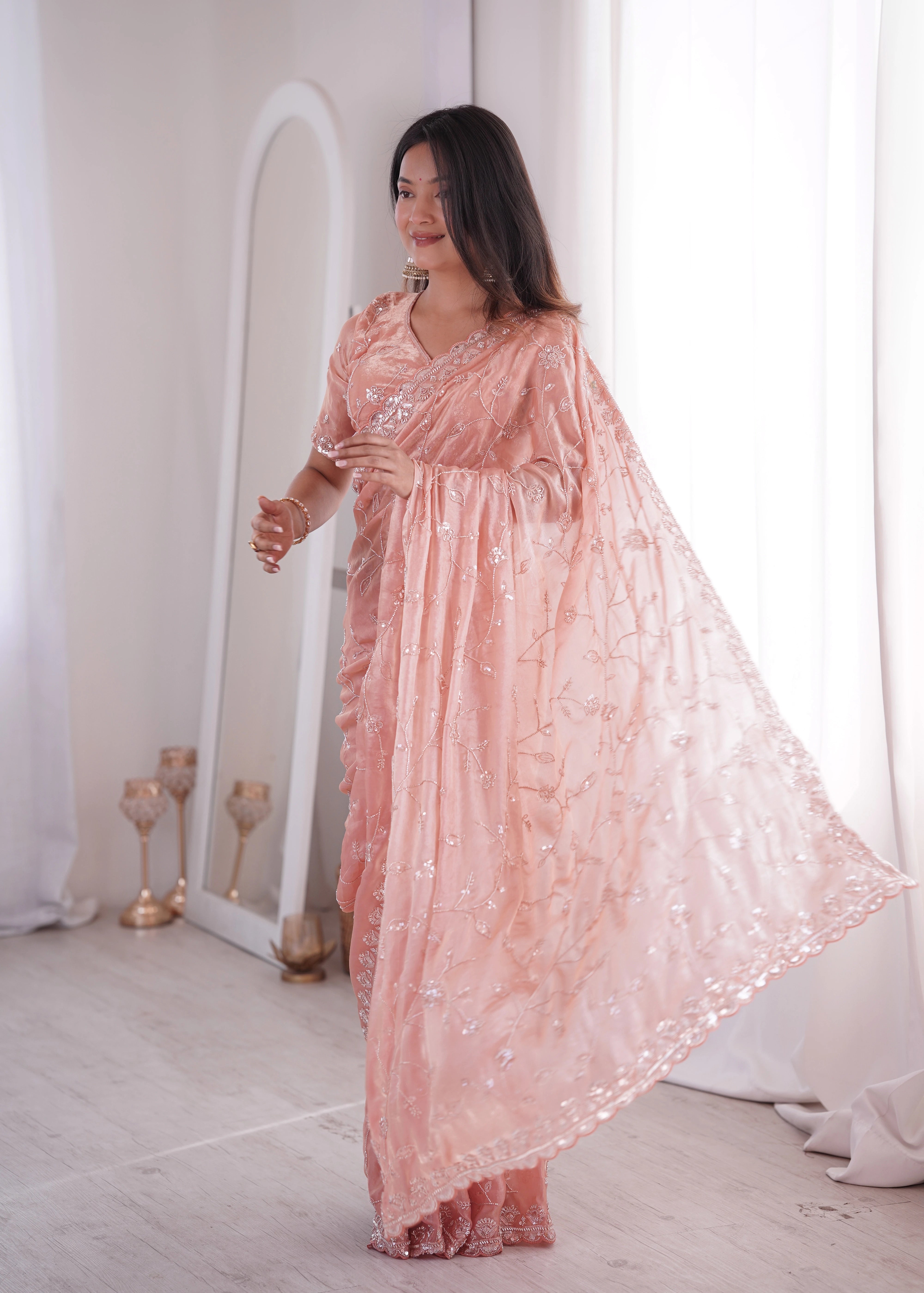Woman in a pink saree standing in a well-lit room with white walls and decor.