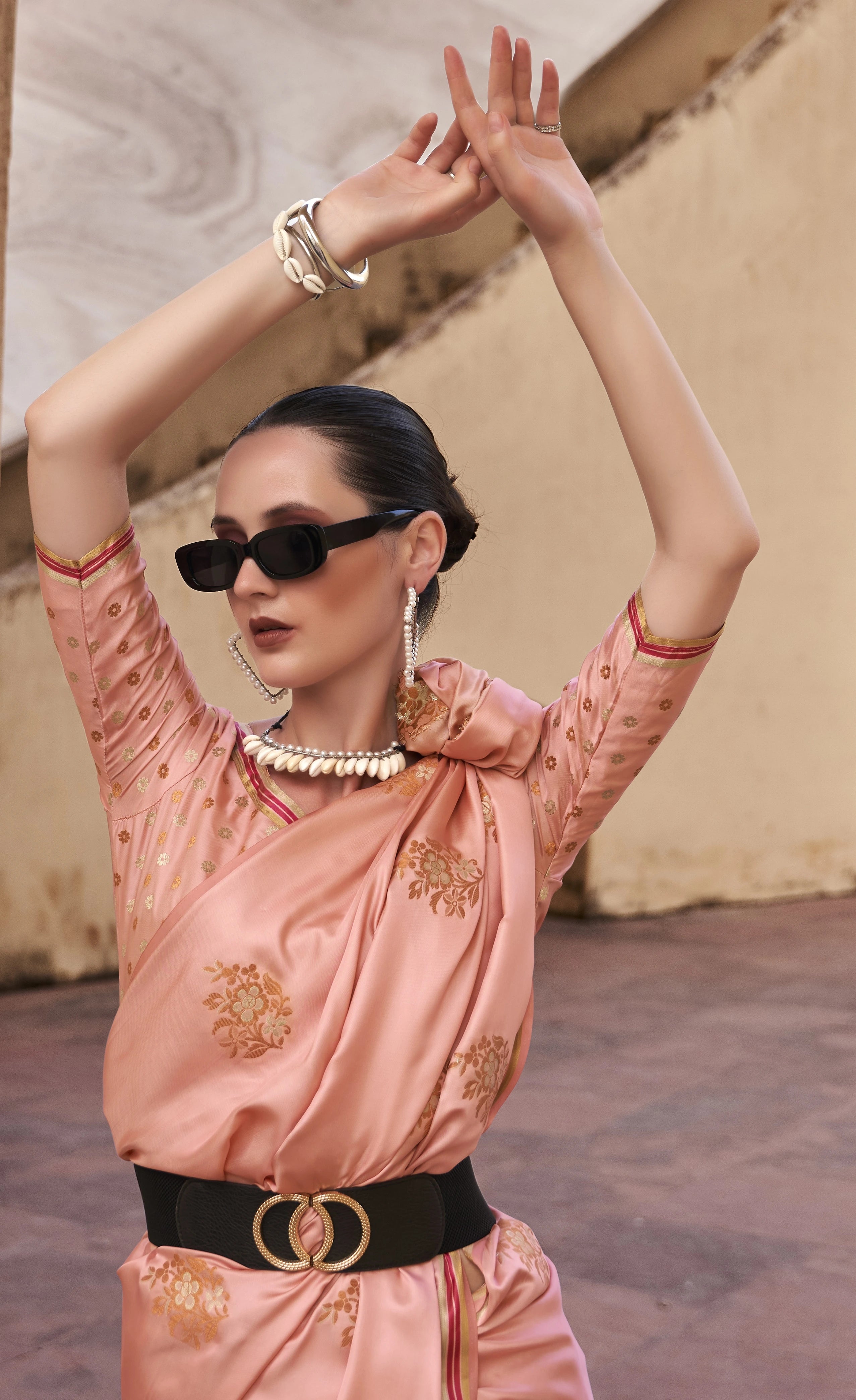 Woman in a pink traditional outfit with gold embroidery, sunglasses, and jewelry, standing against a textured wall.