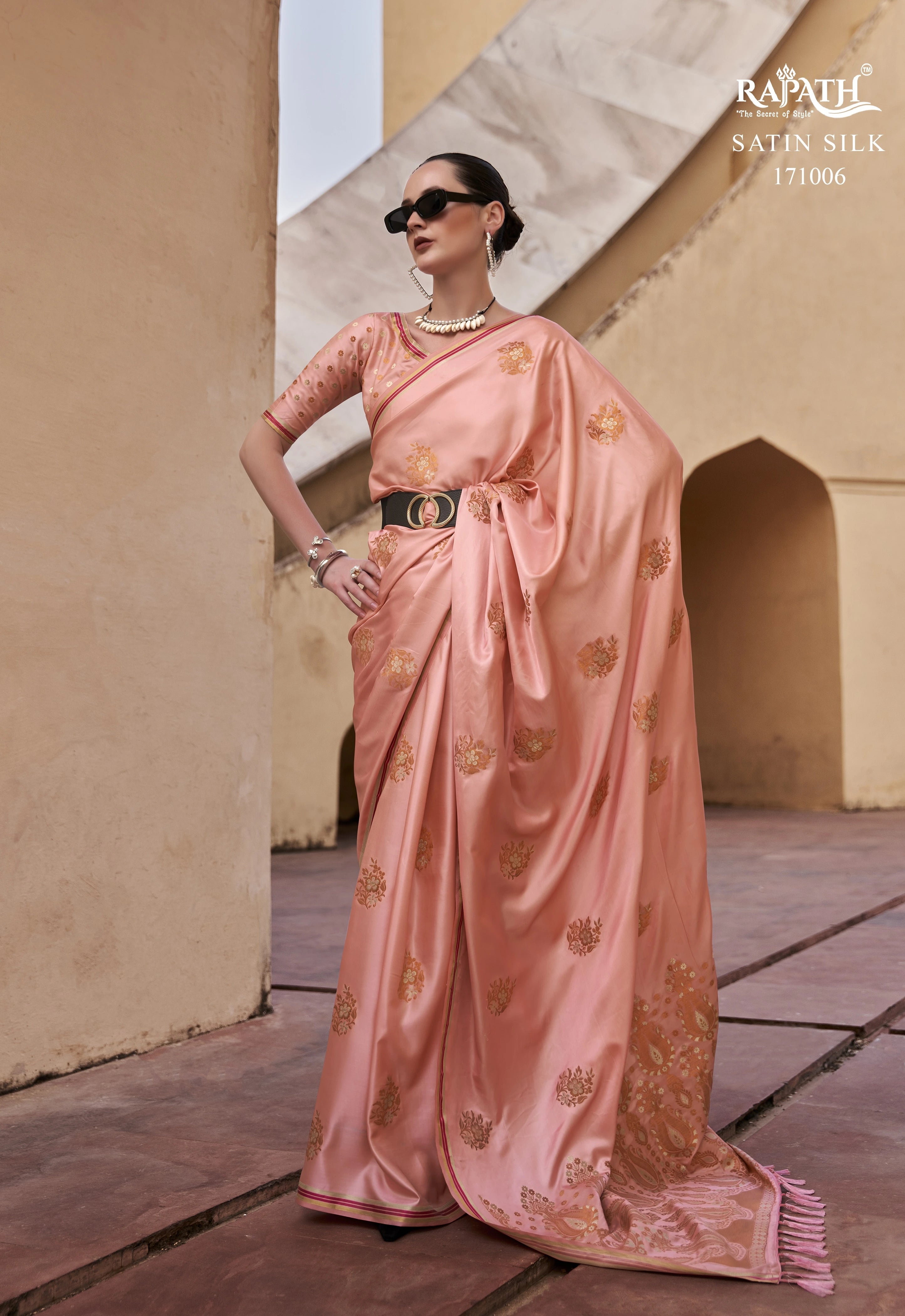 Woman wearing a pink saree with gold patterns in an outdoor setting, with 'Rapun' branding.