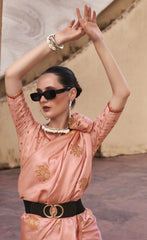 Woman in a pink traditional outfit with gold embroidery, sunglasses, and jewelry, standing against a textured wall.