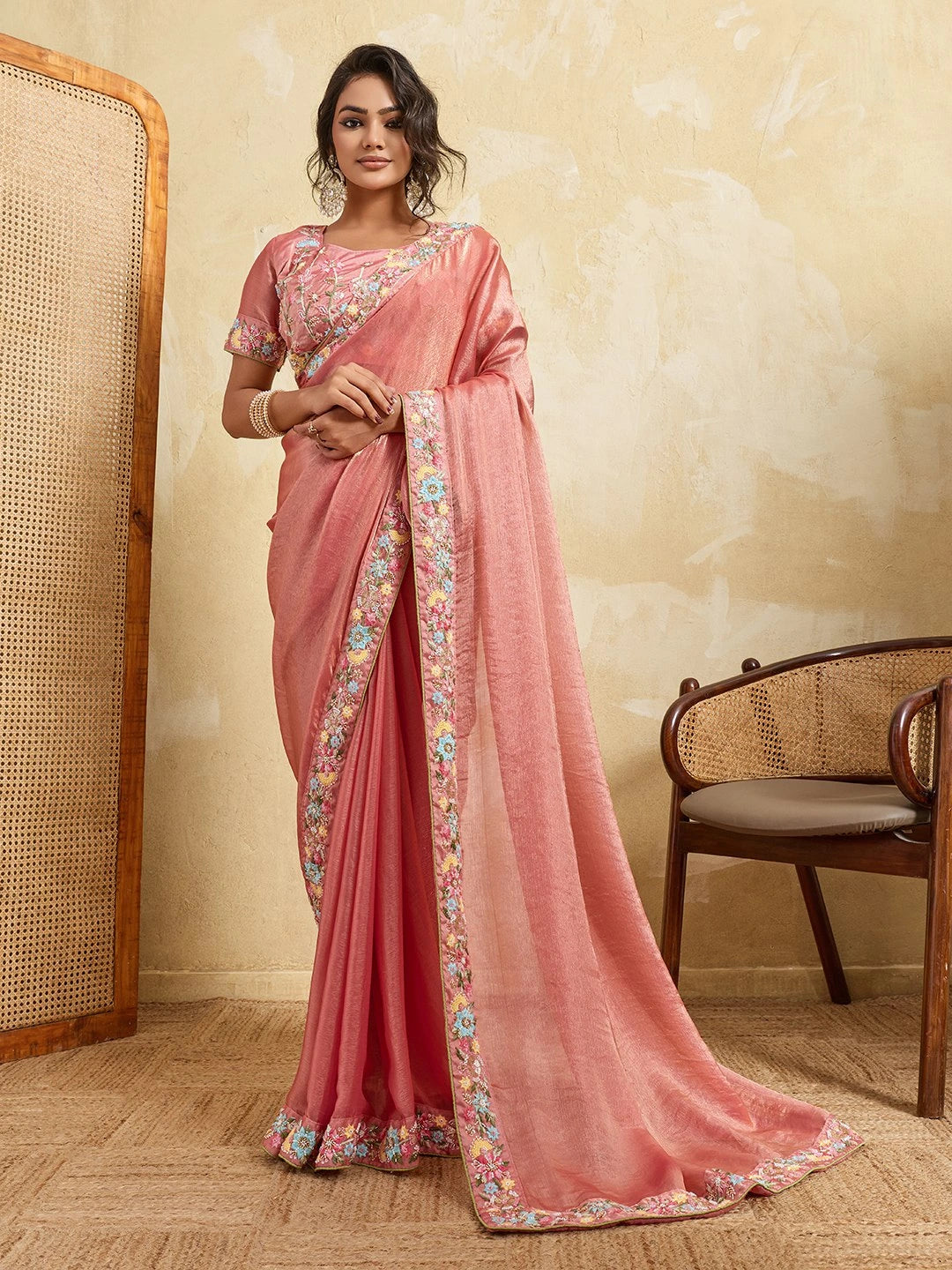 Woman wearing a pink saree with floral patterns in a room with a textured wall and wooden chair.