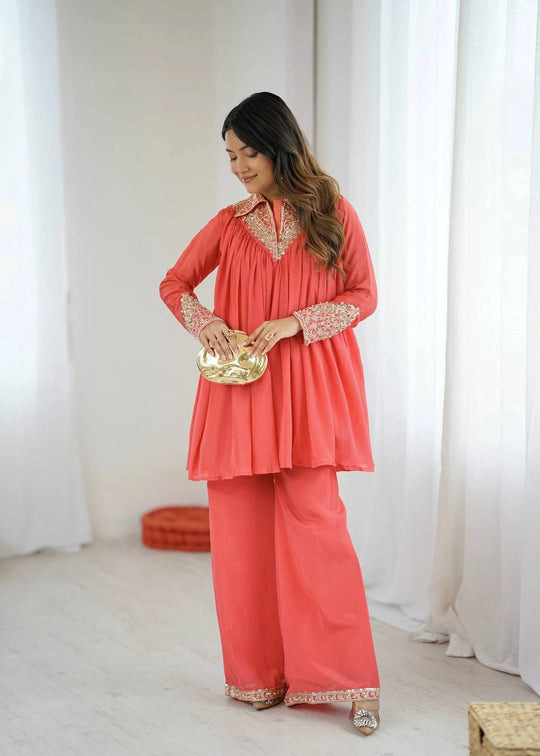 Woman in a coral outfit with gold details holding a gold clutch against a white curtain background